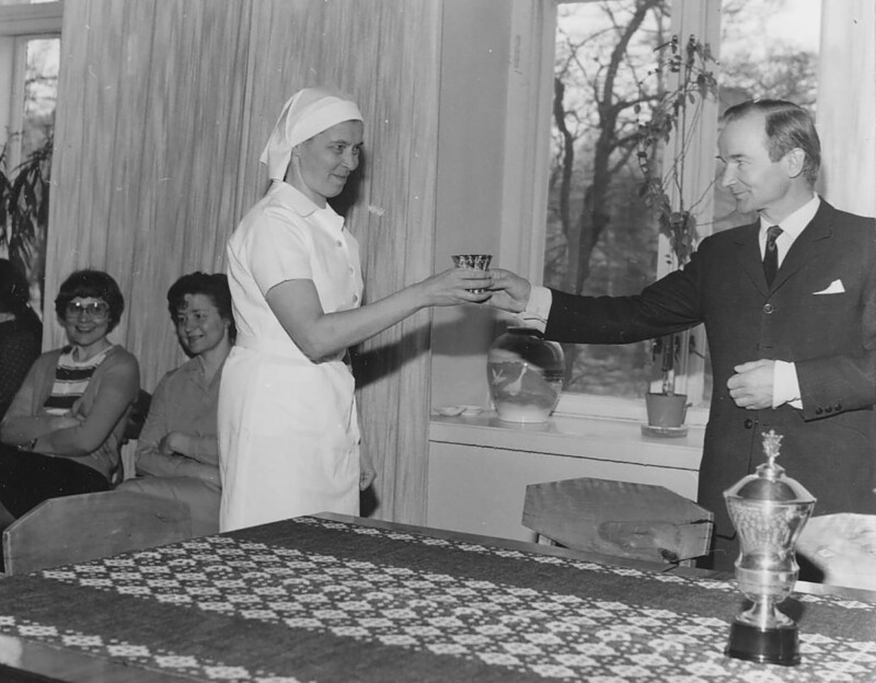 Mustavalkoinen valokuva 1970-luvulta, jossa mies luovuttaa palkinnon hymyilevälle naiselle, joka on pukeutunut hoitajan asuun; taustalla kolme istuvaa henkilöä seuraamassa tilaisuutta.