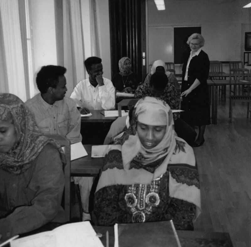 Mustavalkoinen kuva, jossa somalialaisia lähihoitajaopiskelijoita istuu pöytien ääressä luokassa, ja valkoinen nainen opettajana seisoo takana.