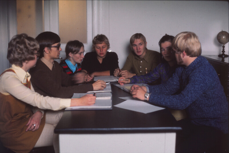 Seitsemän henkilöä istuu pöydän ympärillä keskustelemassa ja lukemassa papereita huoneessa vuonna 1972.