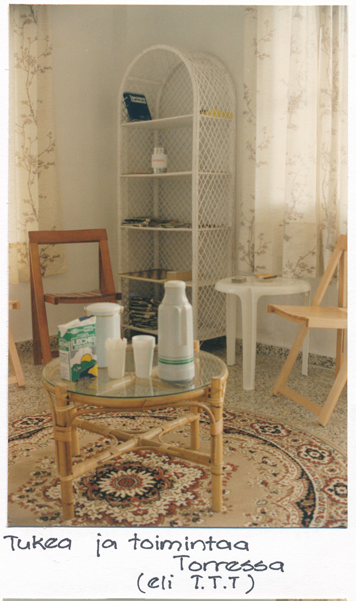 Kuva 1990-luvun diakonissalaitoksen Torren tuvasta, jossa näkyy keinutuoli, pyöreä lasipöytä, valkoinen hyllykkö ja kukkakuvioitu matto, pöydällä termospullo, maitopurkki ja muovimukeja.