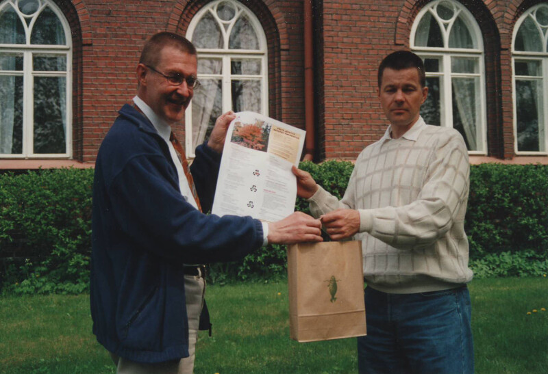 Kaksi miestä ulkona, taustalla punatiilinen rakennus ja ikkunat, toinen heistä pitää kädessään paperidokumenttia ja toinen ruskeaa paperipussia.