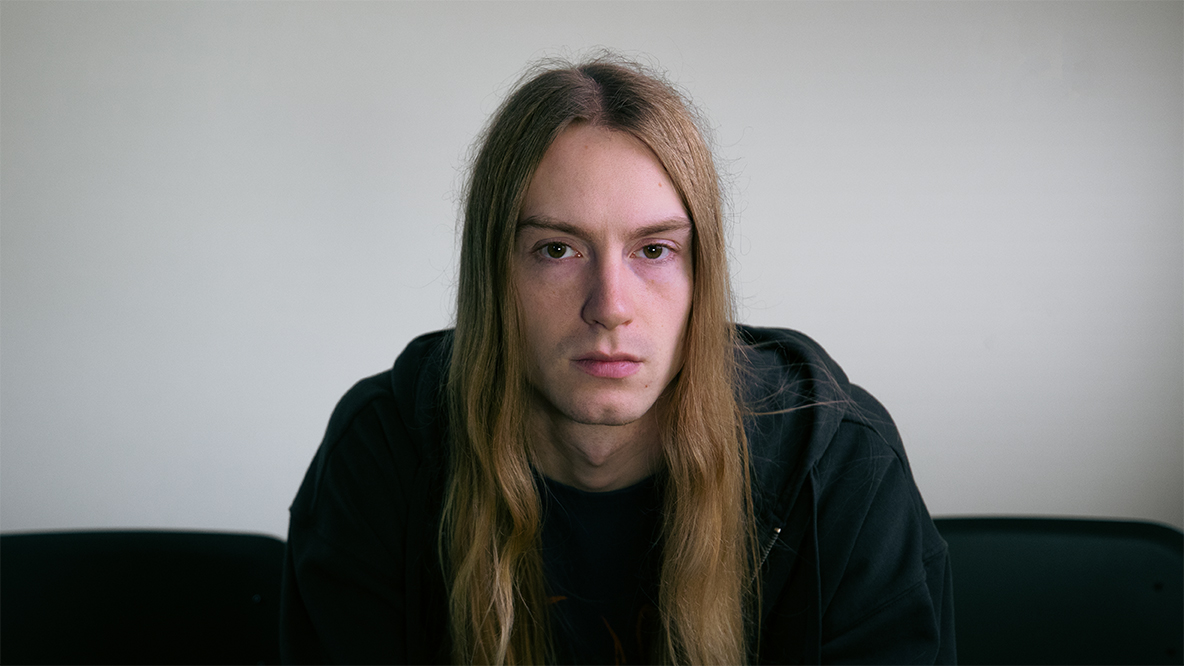 Nuori pitkähiuksinen henkilö katsoo suoraan kameraan vakavana, taustalla harmaa seinä.