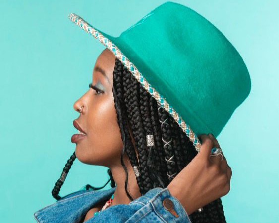 Profiilikuva tummaihoisesta naisesta, joka katsoo vasemmalle; hänellä on pitkät letit, nenäkoru ja sormuksia sekä kirkkaanvihreä huopahattu, taustana vaaleanturkoosi.