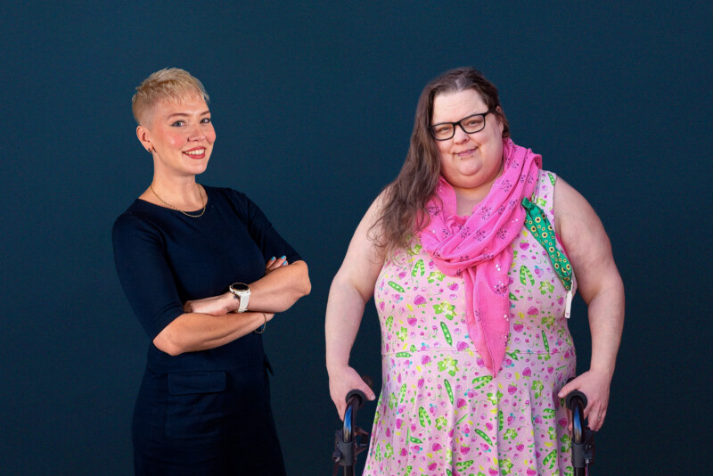 Kuvassa kaksi hymyilevää henkilöä tummansinistä taustaa vasten: vasemmalla lyhythiuksinen henkilö tummissa vaatteissa seisoo käsivarret puuskassa, oikealla kukkamekko ja vaaleanpunainen huivi päällä oleva henkilö nojaa kävelytukeen.