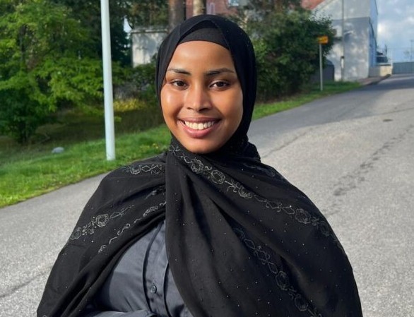 Hymyilevä nuori nainen mustassa huivissa ja mustassa viitassa harmaan paidan päällä seisoo ulkona kävelytiellä, taustalla vihreää kasvillisuutta ja rakennuksia.