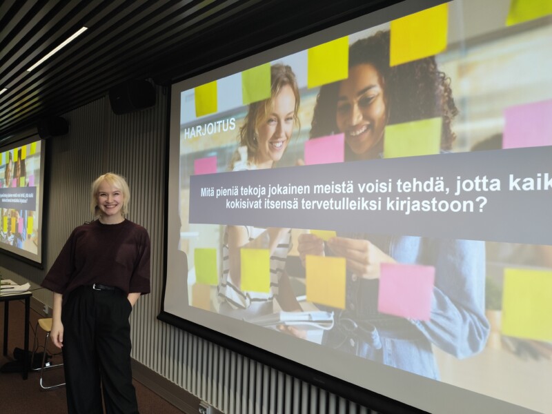 Esiintyjä hymyilee seisten luentosalin edessä valkokankaan vieressä, jossa näkyy esitysdia kysymyksellä "Mitä pieniä tekoja jokainen meistä voisi tehdä, jotta kaikki kokisivat itsensä tervetulleiksi kirjastoon?" ja värikkäitä muistilappuja.