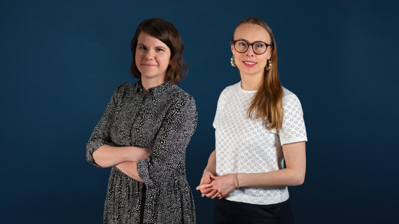 Kaksi hymyilevää naista seisoo tummansinisen taustan edessä; vasemmalla ruskeat puolipitkät hiukset ja kuvioitu pitkähihaismekko, kädet ristissä, oikealla pitkä vaalea hius, pyöreät silmälasit ja valkoinen lyhythihainen paita, kädet edessä.