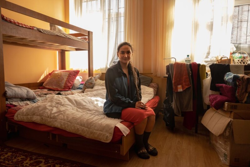 Nainen istuu sängyllä, jonka vieressä on tavaraa laatikoissa ja tuolin karmeilla.