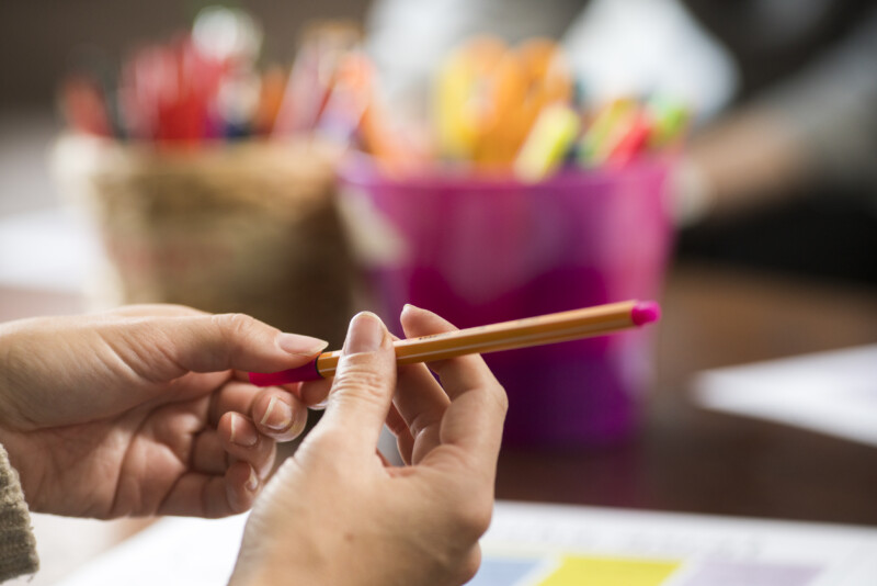 Lähikuva käsistä, jotka pitelevät lyijykynää paperin päällä, taustalla sumeina värikkäitä kynäpurkkeja pöydällä