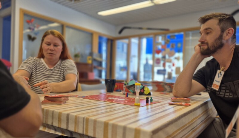 Henkilöt istuvat pöydän ääressä pelaamassa Aliasta.