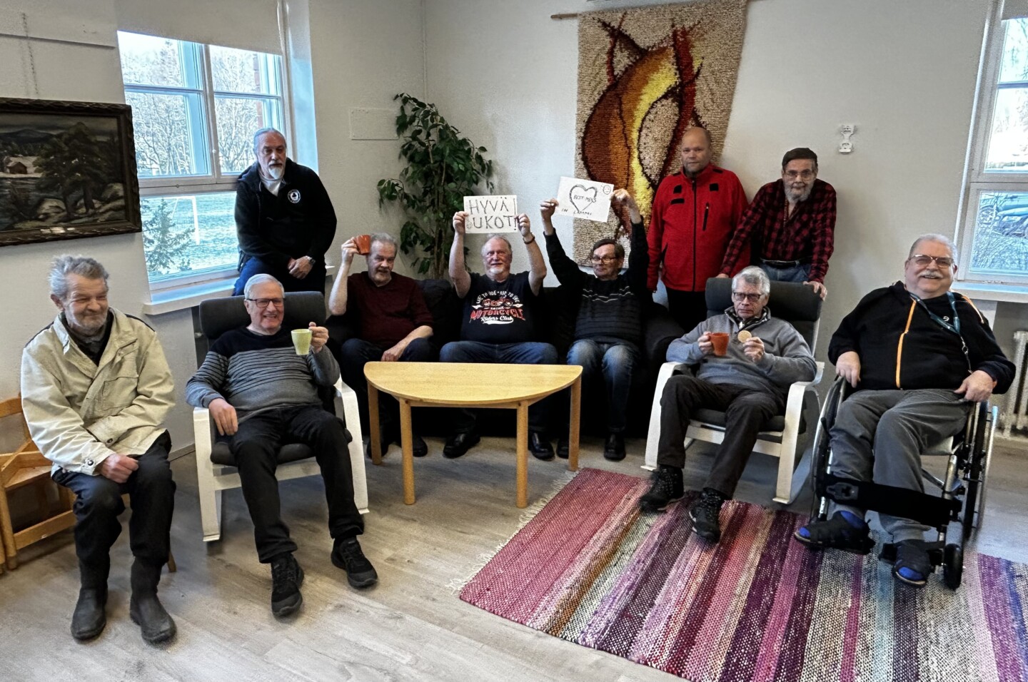Ihmisiä istumassa tuoleilla ja sohvalla, osalla heistä on kylttejä joissa toivotetaan hyvää miestenpäivää