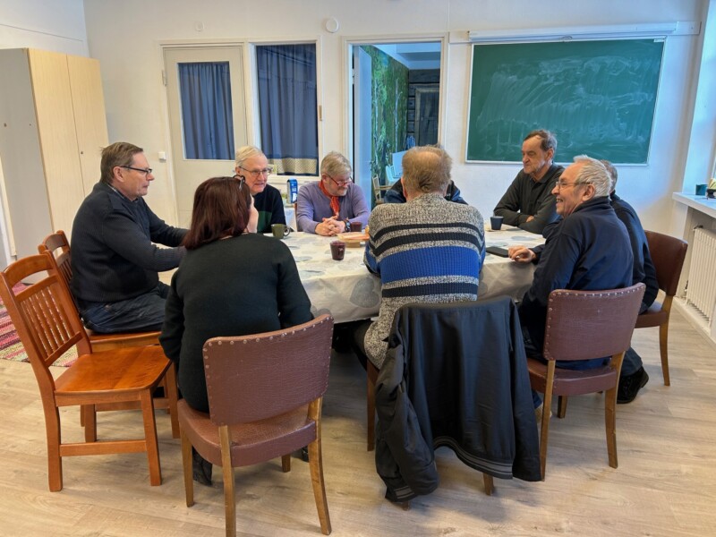 Yhdeksän henkilöä istuu pöydän äärellä, osa hymyilee