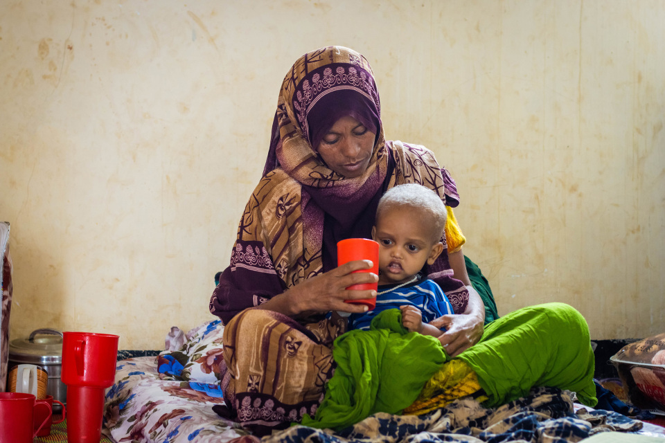 Nainen huivissa istuu sängyllä sylissään pieni lapsi, joka juo punaisesta mukista — intiimi hoivahetki yksinkertaisessa huoneessa.