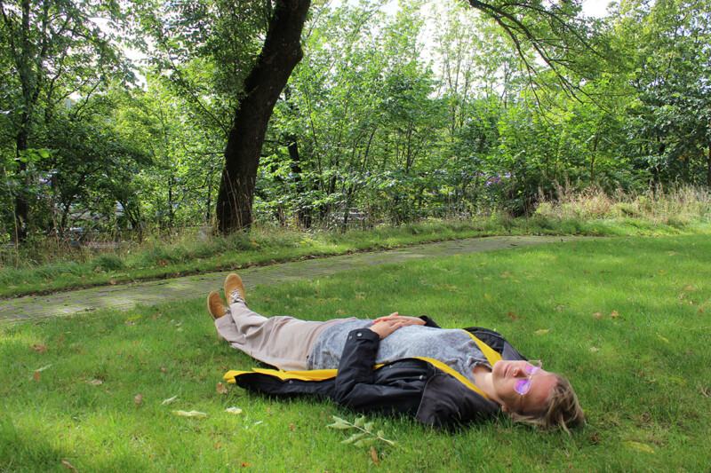 Henkilö makaa nurmikolla puiston rehevässä ympäristössä aurinkolaseissa ja nahkatakki päällä, kädet ristissä vatsalla, taustalla polku ja lehtipuita.