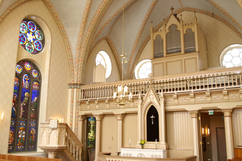 Sisäkuva kirkon salista: vaaleaksi maalattu koristeellinen alttari ristillä, yläpuolella urut ja parvi, vasemmalla korkeat värikkäät lasi-ikkunat ja saarnatuoli, holvikatto kullankeltaisin koristein.