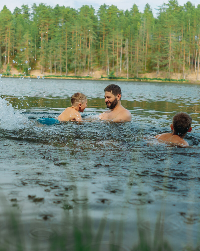 Aikuinen mies ja kaksi lasta uivat ja leikkivät järvessä, taustalla metsää.