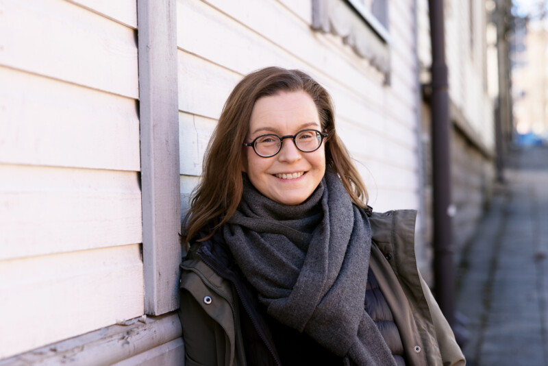 Hymyilevä nainen silmälasit päässä nojaa valkoisen puutalon seinään Vallilan kadulla, pukeutuneena tummaan takkiin ja paksuun harmaaseen huiviin.