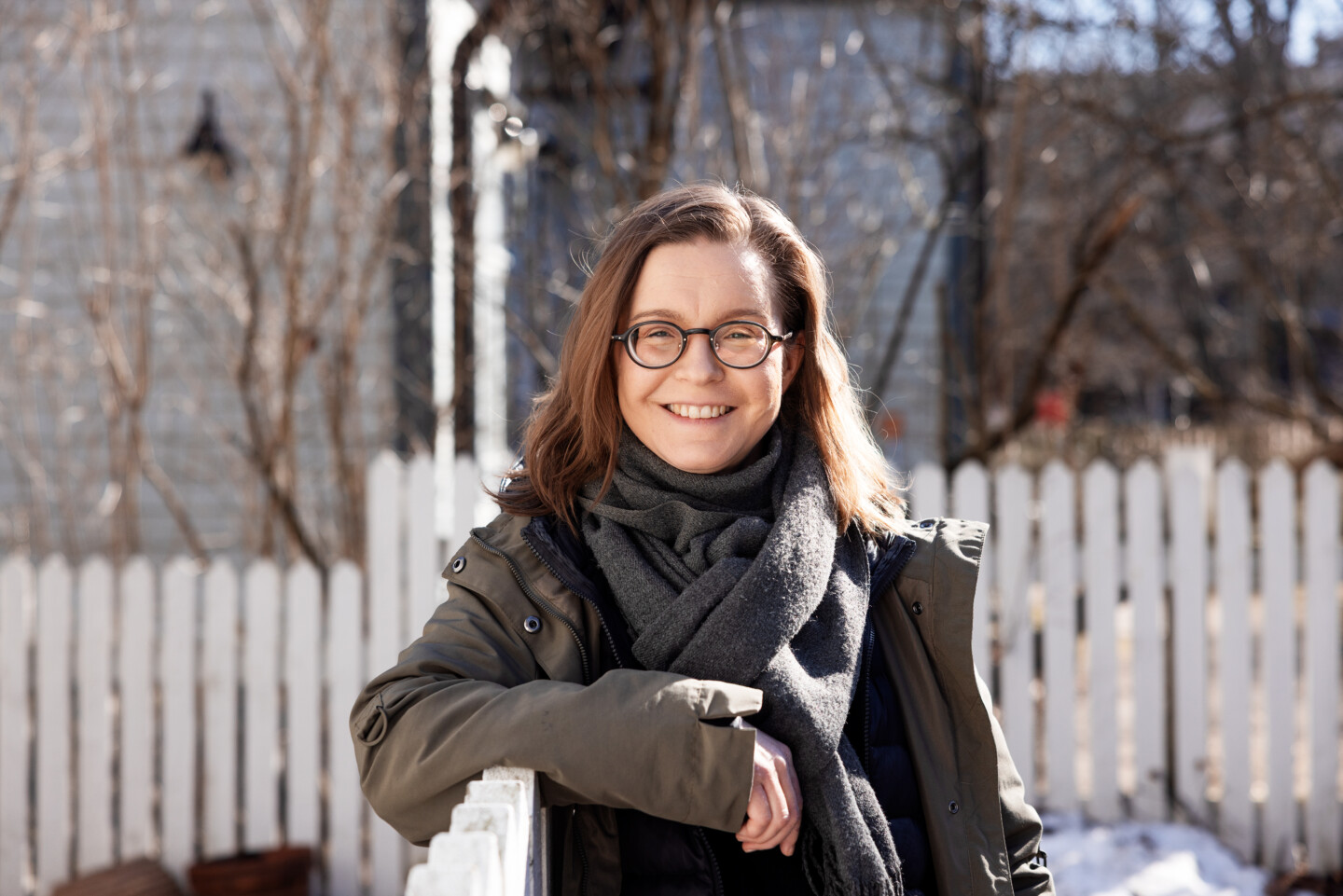 Ulkoilma muotokuva hymyilevästä naisesta silmälasit päässä, ruskeat olkapituisiin ulottuvat hiukset, tumma talvitakki ja huivi, nojaa valkoiseen aitaan, taustalla lumipeitteinen piha, lehdettömiä puita ja puutalo.