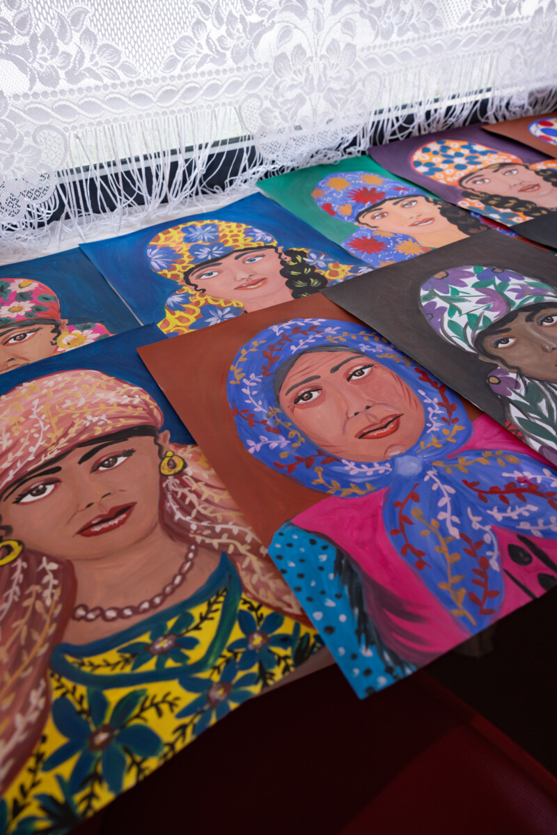 Useita maalauksia romaninaisista, värikkäät kukkakuviolliset huivit, korut ja kirjavat vaatteet korostettuina eri kasvonpiirteillä; teokset on aseteltu vierekkäin pöydälle pitsiverhon alla.