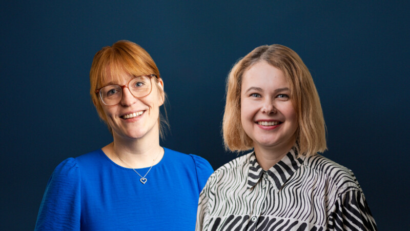 Kaksi hymyilevää aikuista naista puolivartalokuvassa sinertävää taustaa vasten; vasemmalla lasit ja sininen pusero, oikealla vaalea polkkatukka ja mustavalkoinen kuvioitu paita.