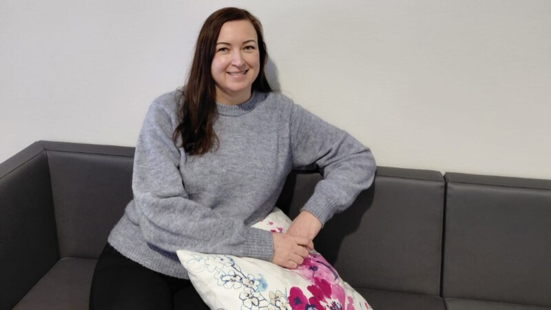 Aikuinen nainen istuu harmaalla sohvalla ja nojaa kukallisella tyynyllä, pukeutuneena vaaleanharmaaseen neuleeseen ja hymyilee vaaleaa seinää vasten.