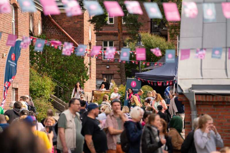Naapuruston ulkotapahtuma tiiliseinien välissä: värikkäitä viirejä roikkuu katoksena, kojuja ja telttoja reunustavat katua ja ihmisjoukko keskustelee ja kävelee etualalla.