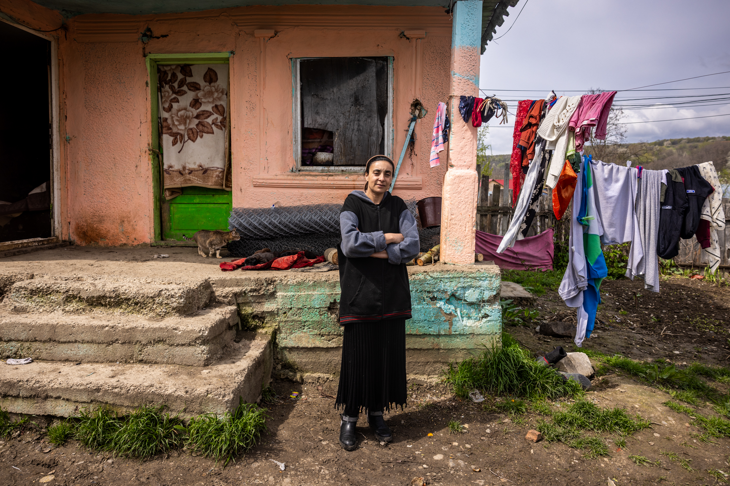A woman stands smiling with her arms crossed on the porch of a weathered peach-colored rural house; concrete steps and clothes drying on a line are visible in the muddy yard.