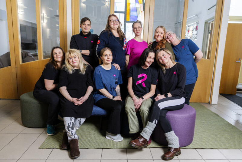 Työryhmä poseeraa rakennuksen sisätilassa: eturivissä viisi istuvaa ja takarivissä kuusi seisovaa henkilöä, kaikki rennosti pukeutuneita ja hymyileviä, taustalla värillinen lasi-ikkuna ja ovia, kuva viestii yhteishengestä ja turvallisuudesta.