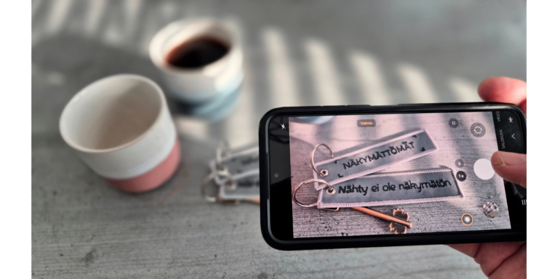 Taustalla on kaksi kahvikuppia, joista on toinen on tyhjä ja toinen täynnä. Edustalla on käsi, joka pitelee puhelimen kameraa ja ottaa lähikuvaa tägeistä, joissa lukee: "Nähty ei ole näkymätön" ja "Näkymättömät".