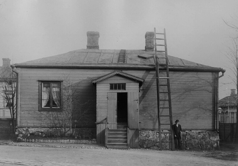 Mustavalkoinen historiallinen valokuva: yksikerroksinen puutalo kivijalalla, keskellä pieni kuisti ja avoin ovi, katolla kaksi savupiippua ja pitkä tikas nojattuna ylös, talon reunalla seisova henkilö, taustalla muita rakennuksia ja paljaita lehtipuita.