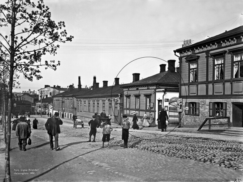Musta‑valkoinen vanha katunäkymä: mukulakivikatu, rivissä puutaloja ja portti oikealla, vasemmalla puu ja käytäviä, useita jalankulkijoita ja lapsia kävelemässä kadulla, taustalla tehtaan piippuja ja kaupunkihorisontti.