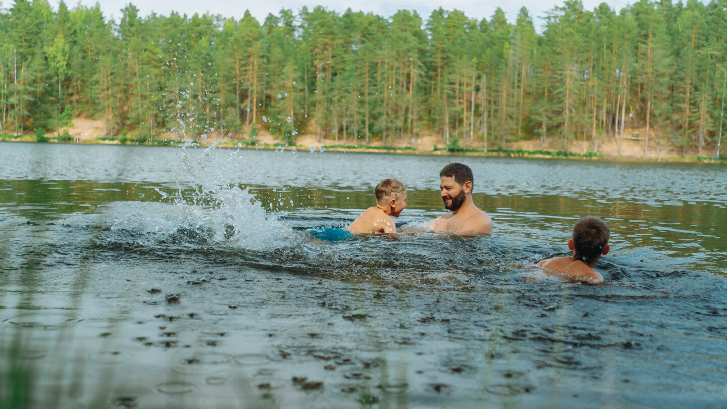 Aikuinen mies ja kaksi lasta uivat metsäisen järven rannassa; aikuinen leikkii ja nostaa yhtä lasta vedestä ja vesi roiskuu ympärillä.