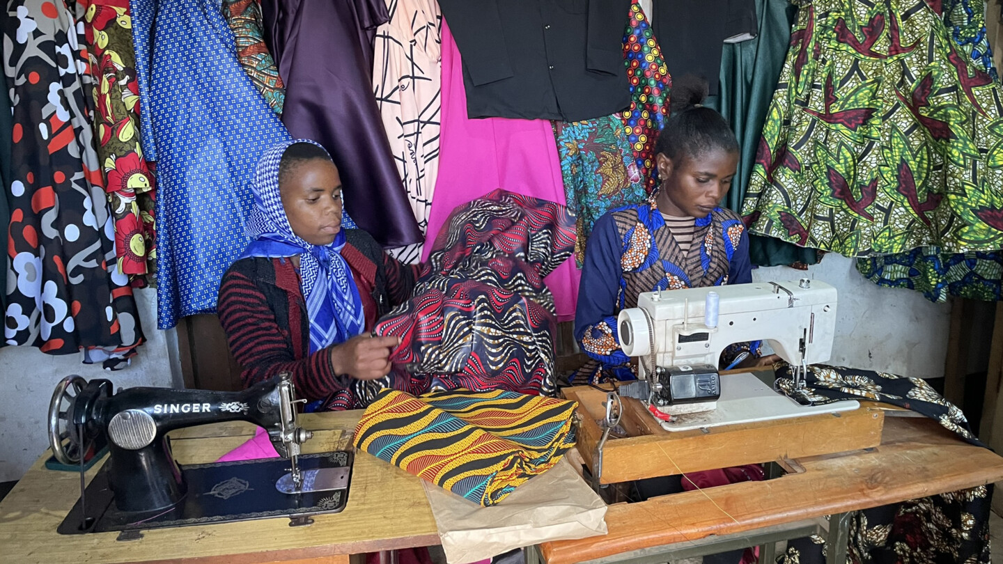 Kaksi naista ompelukoneiden ääressä työpöydän äärellä, pöydällä ja taustalla värikkäitä kuvioituja kankaita ja valmiita kangaspaloja.