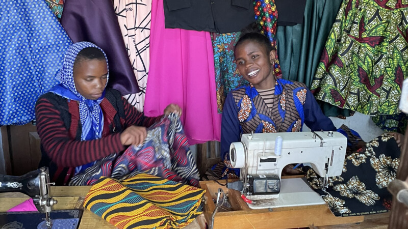 Kaksi naista istuu ompelutyöpöydän ääressä, toinen käyttää ompelukonetta hymyillen ja toinen käsittelee värikästä kangasta, taustalla roikkuu kirkkaita kankaita ja valmiita vaatteita.