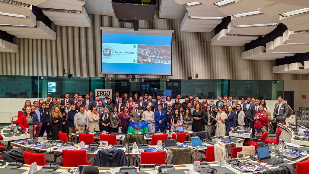 Wide group photo in a large modern conference room showing around 80 people standing behind rows of desks with red chairs and laptops, a presentation screen at the front, and attendees holding a colorful Romani flag.