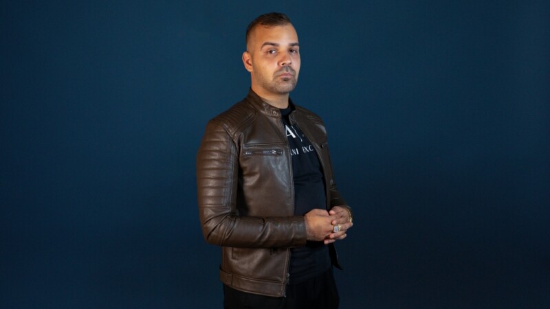 Ruskeaan nahkatakkiin pukeutunut lyhythiuksinen mies seisoo tummansinisen taustan edessä ja katsoo kameraan.