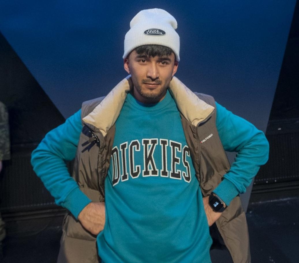 Nuori mies katsoo kameraan, valkoinen pipo päässä, turkoosi DICKIES-collegepaita ja ruskeahko toppaliivi yllä, kädet lanteilla tummansinisellä näyttämötaustalla.