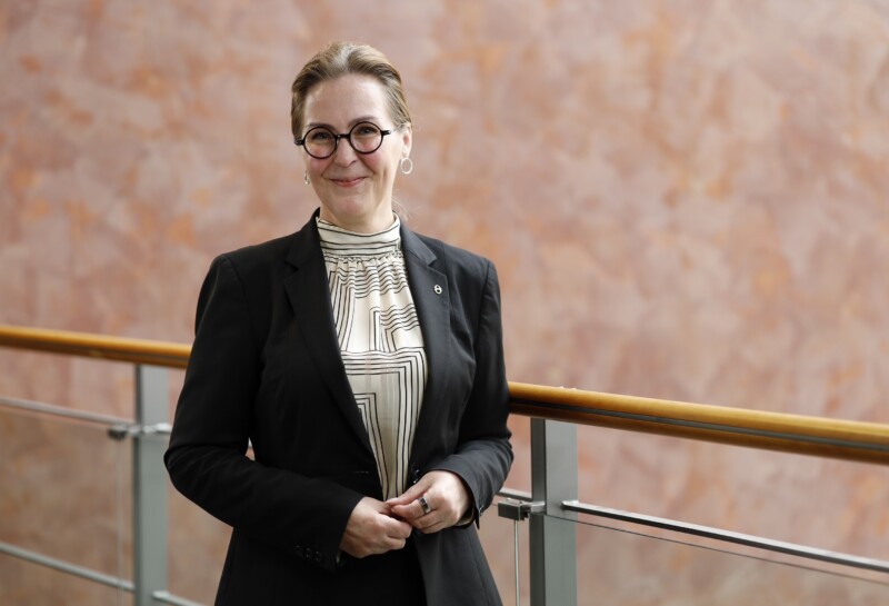 Hymyilevä nainen laseissa ja tummassa bleiserissä seisoo sisätilan lasikaiteen vieressä kädet yhteen kiedottuina vaalean marmoriseinän edessä.