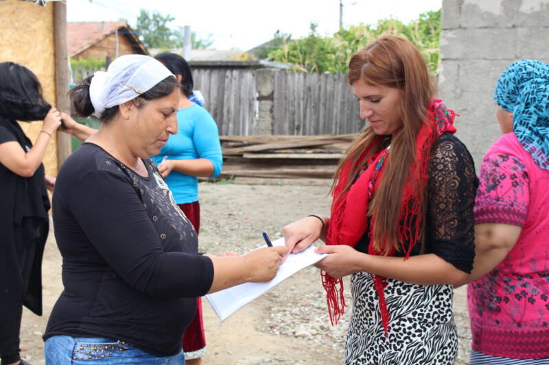 Huivipäinen nainen allekirjoittaa toisen naisen pitelemää paperia ulkona.