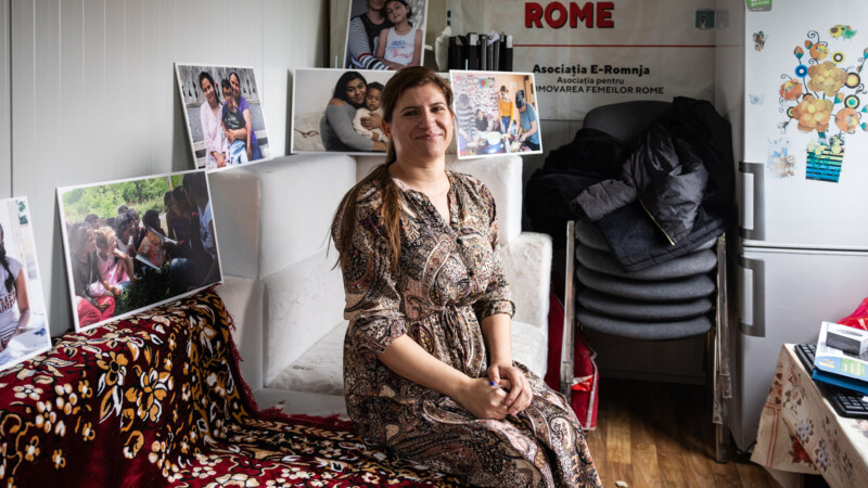 Hymyilevä nainen istuu sohvalla kukkakuvioinen mekko yllä, ympärillä esillä valokuvia ja arjen tavaroita sekä pinottuja tuoleja.