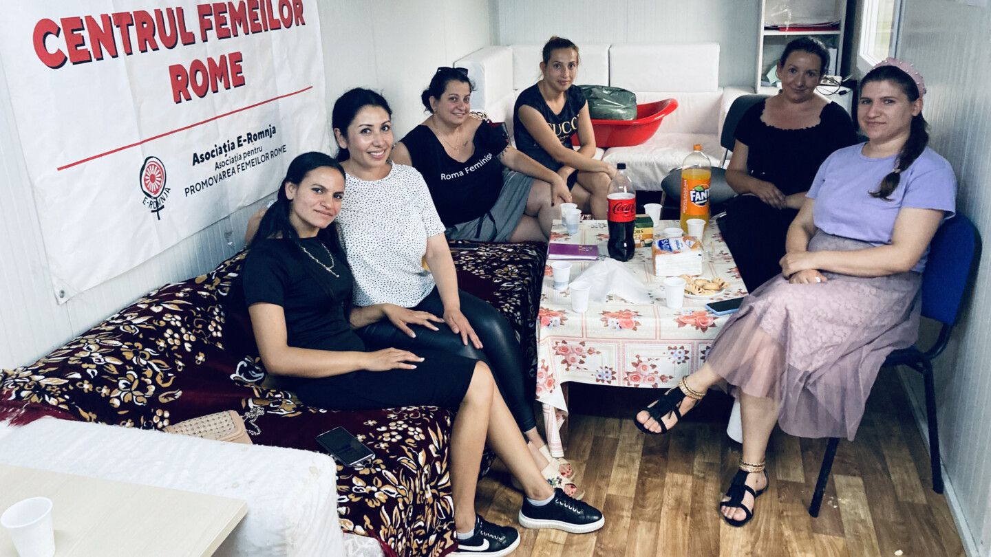 Kuusi hymyilevää naista istuu huoneen pöydän ympärillä olevalla sohvalla ja tuoleilla, pöydällä juomia ja pikkupurtavaa.