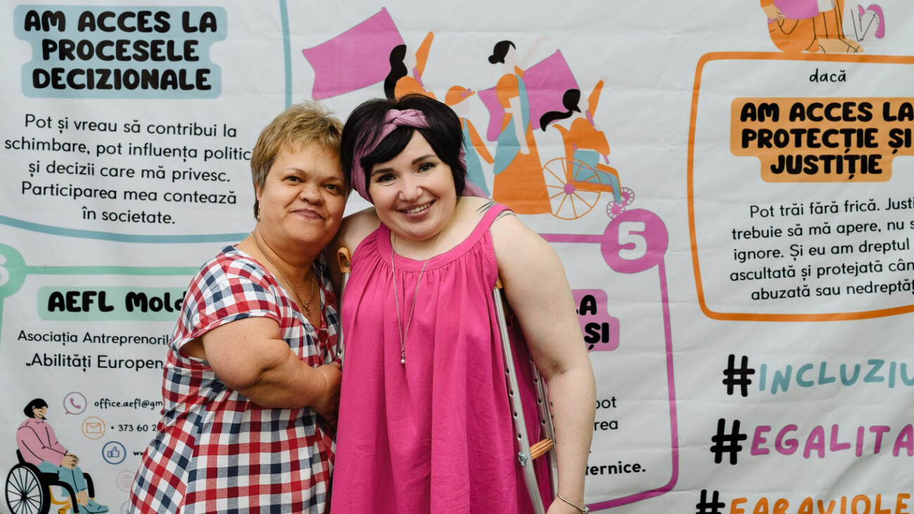 Kaksi hymyilevää naista halaavat toisiaan tapahtuman julisteiden edessä; toisella on ruudullinen paita ja toisella pinkki hiuspanta ja mekko, taustalla värikkäitä julisteita.