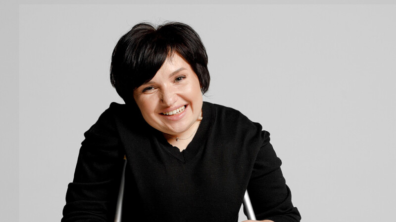 Hymyilevä tummahiuksinen nainen mustassa neuleessa nojautuu kävelykeppeihinsä ja katsoo suoraan kameraan vaaleaa taustaa vasten.