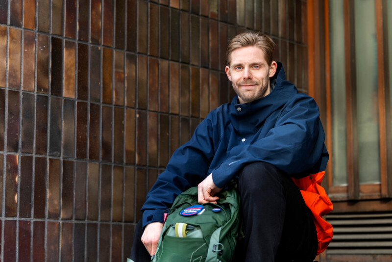 Kuvassa sinitakkinen mies istuu rakennuksen ulko-oven edessä. Hänellä on vihreä reppu sylissä ja hän hymyilee lempeästi kameralle.