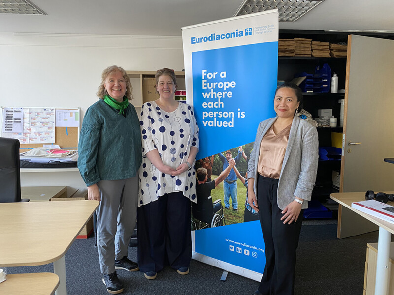 Kolme naista seisoo toimistoympäristössä hymyillen Eurodiaconia-rollupin vieressä.