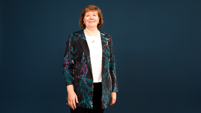 Asiantuntija kuva, jossa hymyilevä nainen seisoo tummansinistä taustaa vasten pukeutuneena valkoiseen paitaan ja tummaan turkoosi–violetin kuvioituun bleiseriin.