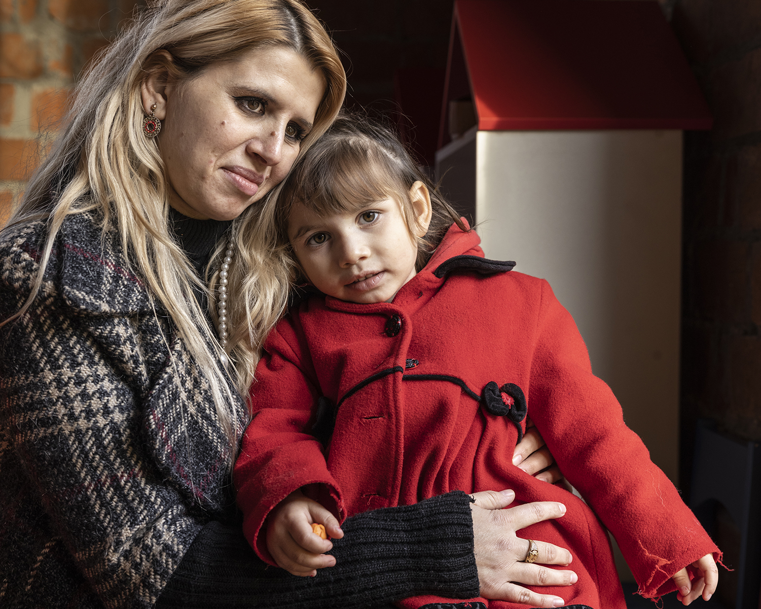 Nainen istuu ja pitää pientä tyttöä käsivarrellaan, tyttö on punaisessa takissa ja pitää pientä oranssia lelua, taustalla tiiliseinä ja hämärä sisätila.