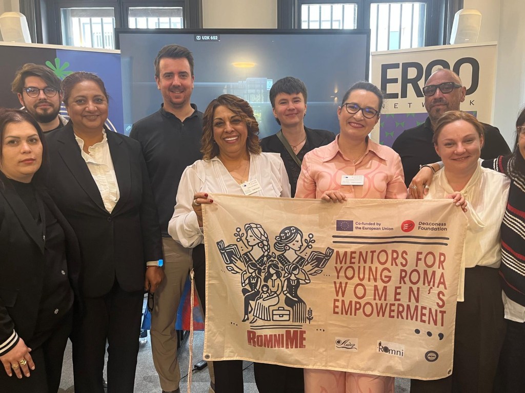 Group of people standing in a conference room smiling and holding a banner reading "Mentors for Young Roma Women's Empowerment".