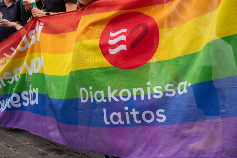 Sateenkaarivärinen pride-lippu, jonka keskellä punainen ympyrälogo valkoisella symbolilla ja teksti "Diakonissalaitos", ihmisten kädet pitelevät lippua katukulkueessa.