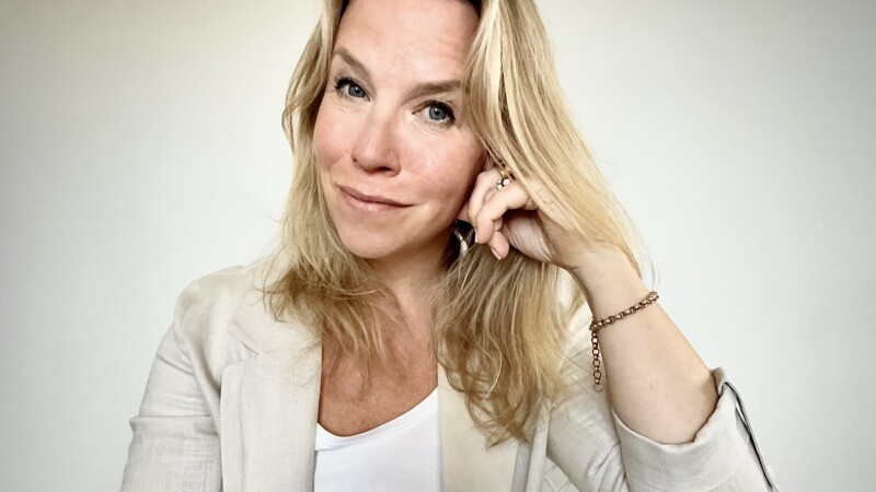 Vaaleahiuksinen nainen hymyilee katsojaan päin ja tukee poskeaan kädellä, pukeutuneena valkoiseen toppiin ja vaaleaan bleiseriin vaalealla taustalla.